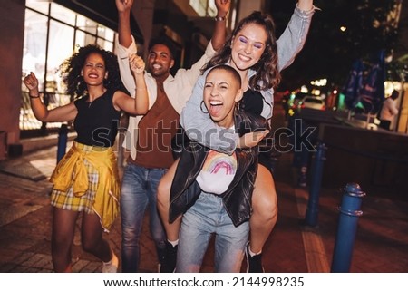 Cheerful young woman piggybacking her friend in the city. Girlfriends cheering and having fun while going out with their friends at night. Group of vibrant friends hanging out together on the weekend. Royalty-Free Stock Photo #2144998235
