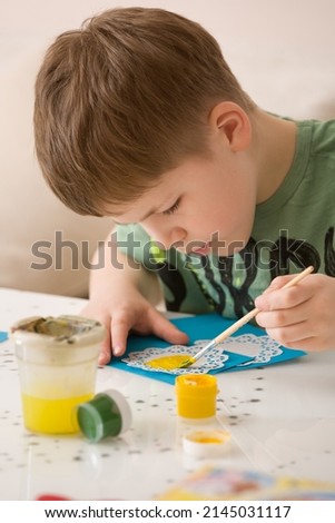 Cute kid boy sitting at the table and drawing with paint. Making crafts at home. Hobby and children activities. Lifestyle concept