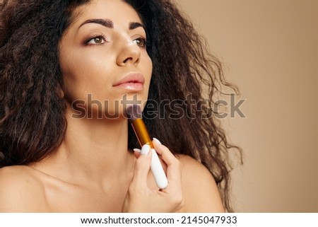 GOOD FOUNDATION CONCEPT. Smiling tanned awesome curly Latin lady hold makeup brush applying foundation with brush after morning shower posing isolated on pastel beige background look aside