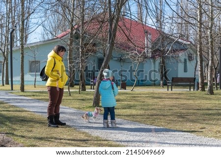 Ukrainian family taking a walk in a spring city park. Mother, five-year-old daughter, and a Chihuahua dog. A turquoise building with a red sloping roof in the background.