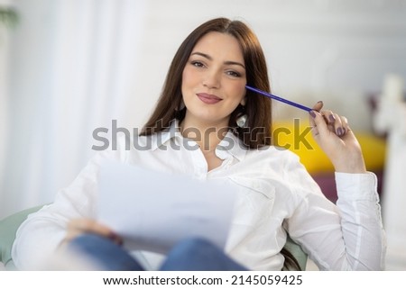 An adult attractive dark-haired girl in a neat stylish white shirt sits near a desk and draws her creative ideas on white paper and in a notebook, in a bright cozy room