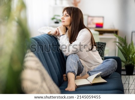 An attractive dark-haired adult girl in a stylish white shirt sits relaxed on a soft blue sofa in her cozy bright apartment, and enthusiastically writes something in her diary