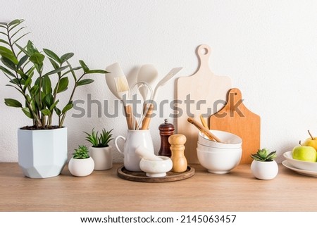 a variety of kitchen utensils, utensils, a potted flower and fruit on a plate on a wooden countertop against a white textured wall. front view Royalty-Free Stock Photo #2145063457
