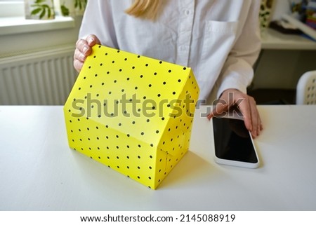 Yellow gift box and smartphone in woman's hands. Gift box, send a gift. Box close-up. Gifts for birthday holidays. Delivery service. Postal services, delivery. Empty packaging, empty space