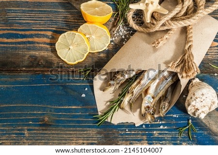 Sun-dried smelt in a paper package. Salted fish with marine decor. Lemon, fresh rosemary, sea rope. Blue nautical wooden background, top view Royalty-Free Stock Photo #2145104007