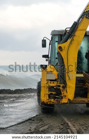 Close-up of a yellow tractor clearing the embankment of sand and mud after a storm against the background of the still raging sea, copy space. Verical photo