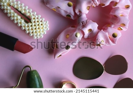 Pink scrunchie, gold earrings and rings, blush, various hair barrettes, lipstick and jade roller on bright pink background. Flat lay.