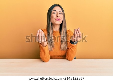 Young hispanic woman wearing casual clothes sitting on the table doing money gesture with hands, asking for salary payment, millionaire business 