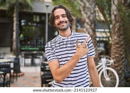 Young hispanic man with beard outdoors at the city cheerful with a smile of face pointing with hand and finger up to the side with happy and natural expression on face 
