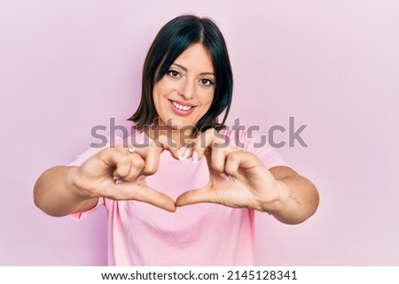 Young hispanic woman wearing casual pink t shirt smiling in love doing heart symbol shape with hands. romantic concept. 