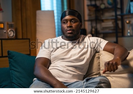 A bored guy is relaxing on the living room sofa in front of the television. The young man stares at a weak movie, TV series, comedies, losing soccer game.