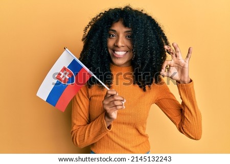 African american woman with afro hair holding slovakia flag doing ok sign with fingers, smiling friendly gesturing excellent symbol 
