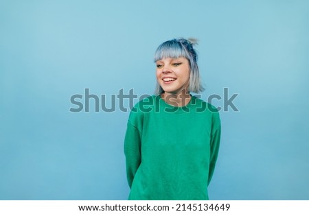 Positive lady with colored hair and in a green sweater stands on a blue background and looks away and laughs.