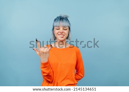 Positive girl in an orange sweatshirt stands on a blue background with closed eyes and shows a gesture of heavy metal with an expressive face. Royalty-Free Stock Photo #2145134651