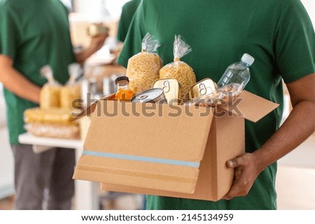 charity, donation and volunteering concept - close up of male volunteer's hands holding box with food over group of people at distribution or refugee assistance center Royalty-Free Stock Photo #2145134959