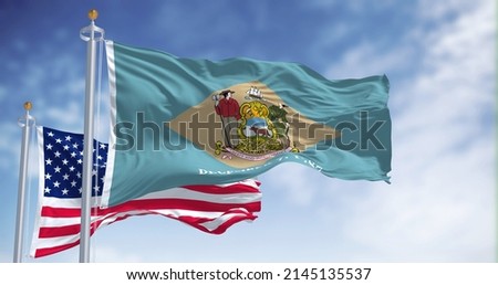 The Delaware state flag waving along with the national flag of the United States of America. In the background there is a clear sky.
