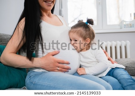 Pretty little girl with ponytails and gold earrings kisses her mother's pregnant belly. Child-bearing caucasian woman hugs her daughter with left hand and toucher tummy with her right hand. They sit.
