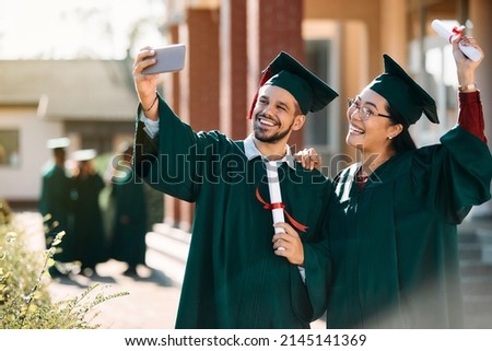 Happy university friends celebrating their graduation and taking selfie with smart phone outdoors.