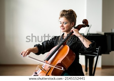 a beautiful girl plays the cello in the classroom against the background of the piano, the cello plays with a bow and smiles, close-up, there is a place for text