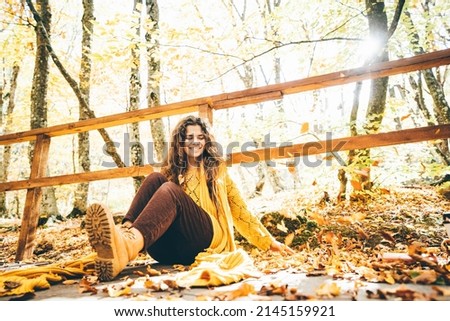 Brunette woman in yellow sweater sitting on a fallen autumn leaves in a park.
