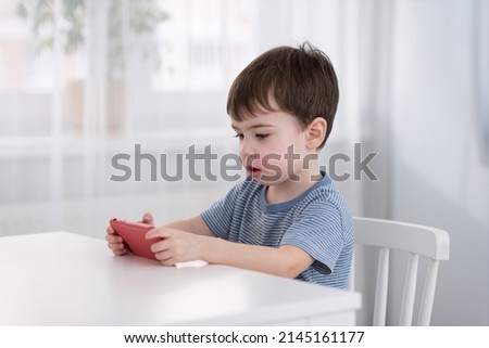 A cute little boy is sitting at the table and playing on the phone. Gadgets and kids concept