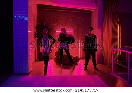 Two teenage girls and guy in posh stage costumes making performance during vogue ball in spacious room or studio lit by neon light Royalty-Free Stock Photo #2145173919