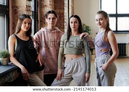 Three teenage girls and guy in activewear standing by window in modern loft studio and looking at camera after vogue dance training Royalty-Free Stock Photo #2145174013