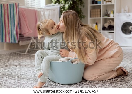 Loving young mom kissing little smiling daughter on cheek, warm family relationships, adorable preschool-aged child spends time with mother in laundry room while doing household chores.