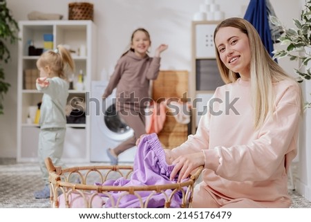 Smiling woman spends time with children while sorting laundry into washing machine, girls dance in background play fooling around, household chores.