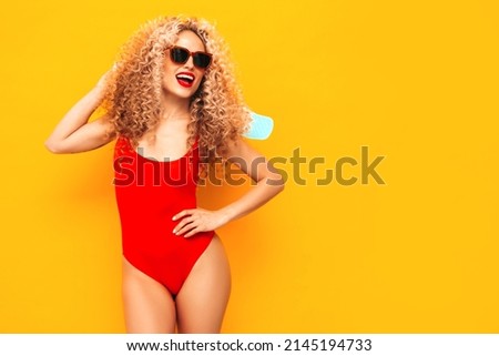 Young beautiful smiling woman posing near yellow wall in studio.Sexy model in red swimwear bathing suit.Positive female with afro curls hairstyle. Holding penny skateboard. Happy and cheerful Royalty-Free Stock Photo #2145194733