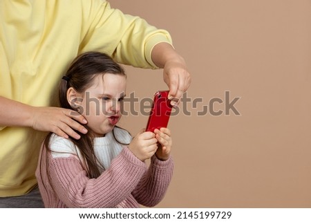 Portrait of parent taking smartphone away from girls hands, beige background. Angry daughter hold tight phone and resist. Concept of prevention of telephone addiction in children, nomophobia. Royalty-Free Stock Photo #2145199729