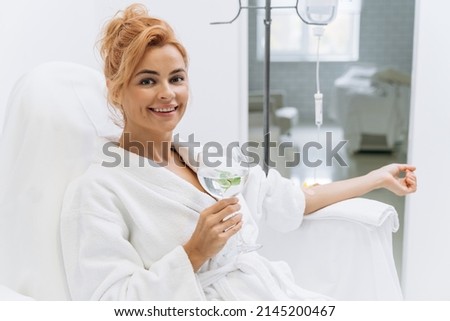 Happy woman in white bathrobe holding glass of lemon beverage and smiling while sitting in armchair and receiving IV infusion Royalty-Free Stock Photo #2145200467