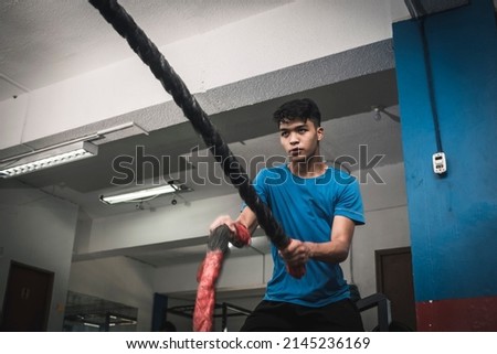 A fit young asian man doing an intense battle rope session. Cardiovascular training at the gym. Royalty-Free Stock Photo #2145236169