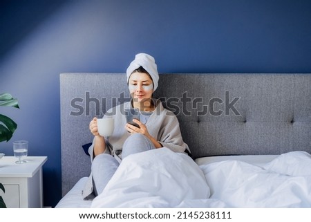 Woman in pajama and bathrobe with eyes patches and towel on her head using phone and drinking morning coffee on her bed. Morning habits and home beauty routine. Work at home. Selective focus. Royalty-Free Stock Photo #2145238111