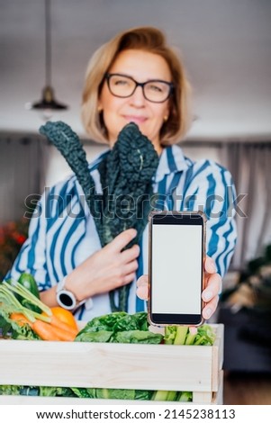 Woman holding phone with blank white screen and fresh kale above wooden box with vegetables. Mockup with copy space for food delivery, diet program, weight loss, recipe box mobile app. Vertical card.