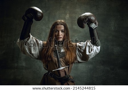 Strength. Brave girl, medieval warrior or knight with dirty wounded face in boxing gloves isolated over dark vintage background. Comparison of eras, history, renaissance style. Fashion, beauty