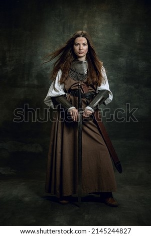 Warlike woman. Portrait of seriuos beautiful woman in image of medieval warrior or knight with dirty wounded face with big sword isolated over dark vintage background. Comparison of eras