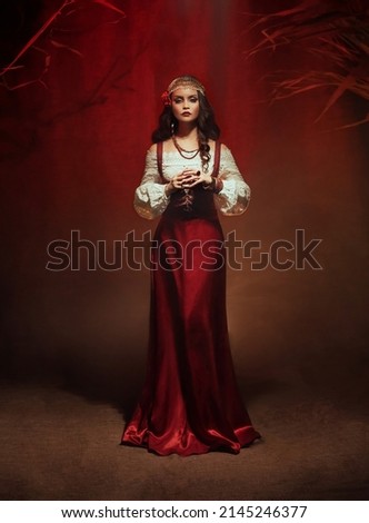  Art photo with noise. Fairy gypsy woman medieval witch stands in dark room. Long black hair, rose hairpin. Red costume vintage dress, fortune teller costume Gold jewelry . Mystical fantasy girl,  Royalty-Free Stock Photo #2145246377
