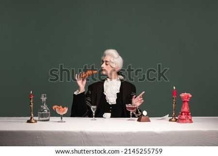 French breakfast. Portrait of young elegant man in peruke and vintage jacket sitting at table isolated on dark green background. Retro style, comparison of eras concept. Royalty-Free Stock Photo #2145255759