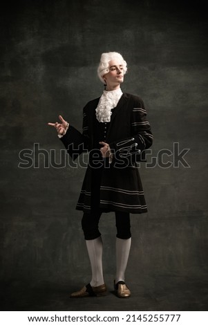 Noble person. Young man wearing wig and vintage medieval outfit like famous composer isolated on dark green vintage background. Retro style, fashion, art, comparison of eras concept. Royalty-Free Stock Photo #2145255777
