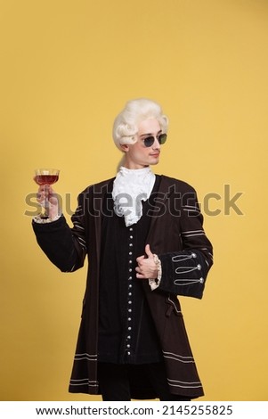Red wine. Portrait of young elegant man in white wig and vintage medieval outfit posing isolated on yellow background. Art, beauty, fashion. Retro style, eras comparison. Copy space for ad Royalty-Free Stock Photo #2145255825