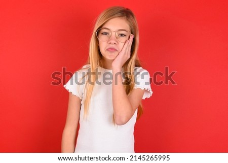Sad lonely blonde little kid girl wearing white t-shirt over red background touches cheek with hand bites lower lip and gazes with displeasure. Bad emotions