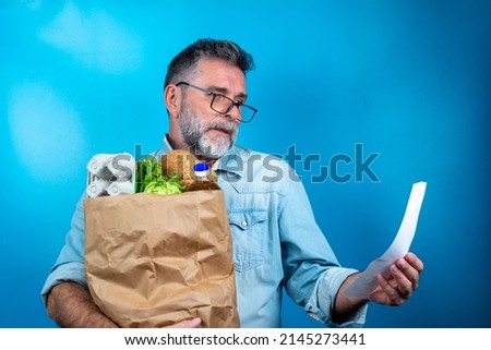 Shocked mature man looking at store receipt after shopping, holding a paper bag with healthy food. Real people expression. Inflation concept. man with a paper bag of groceries looks surprised. Royalty-Free Stock Photo #2145273441