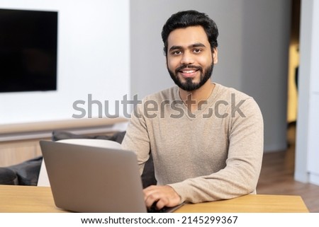 Cheerful Indian man wearing casual wear sitting at the table and using laptop at modern home office, glad ethnic latin male entrepreneur typing on keyboard Royalty-Free Stock Photo #2145299367