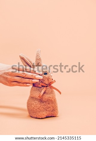 A creative and unusual Easter concept. Female hands hide the eyes of a small puffy brown Easter bunny. Realistic aesthetic look. Contemporary style. Blazing Orange background, soft shadows.