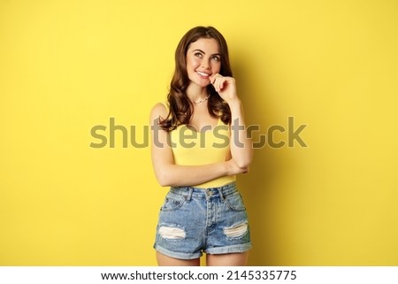 Thinking woman smiling, looking up and deciding, imaging smth, picturing or daydreaming, posing in summer clothes against yellow background
