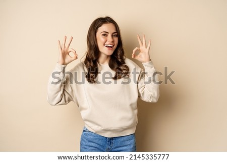 Very good, nice work. Cheerful young woman agrees, shows okay, ok praise, recommending smth, standing over beige background