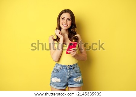 Technology and cellphone. Beautiful stylish girl order in online store, praying in application, using smartphone app and thinking, making decision, standing over yellow background