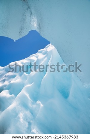 Looking outside of a glacier ice cave into a deep blue sky with tiny drops of water ready to drop from the ceiling with fancy and beautiful shapes of ice in the center of the picture