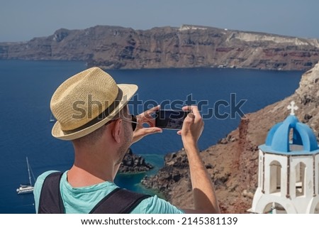 Man in a hat taking picure of classic view of Oia village on Santorini island, Greece, view from the back
